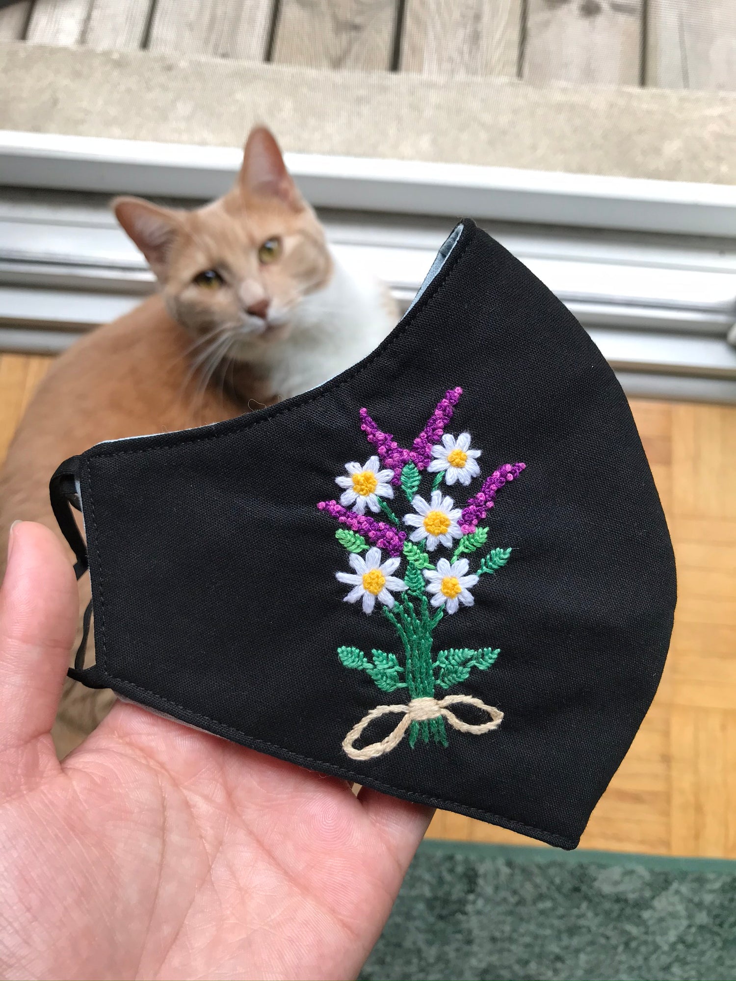 hand holding a black mask embroidered flower pattern with a orange cat in the distance